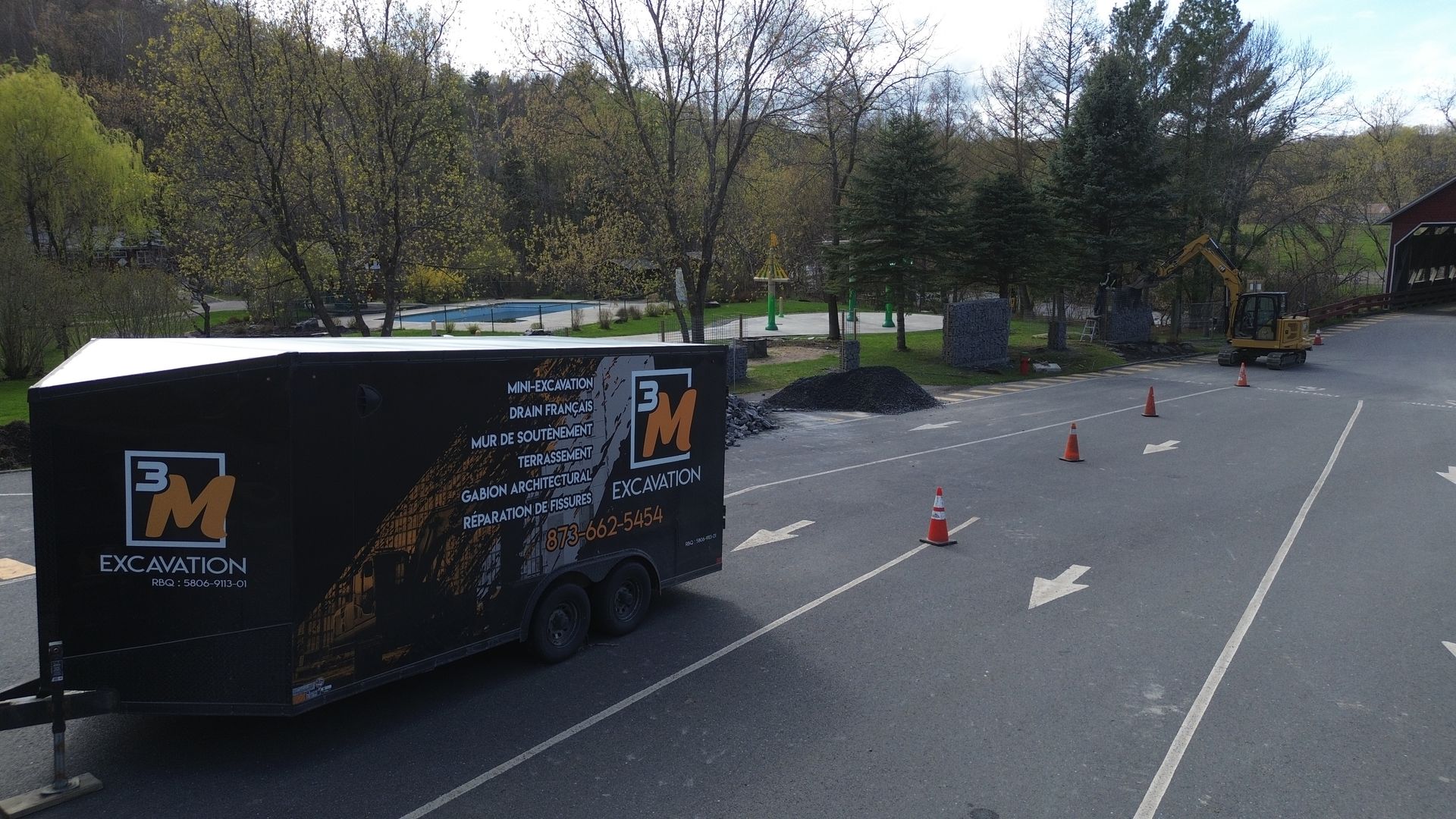 Remorque noire avec logo sur route asphaltée, arbres et cônes.