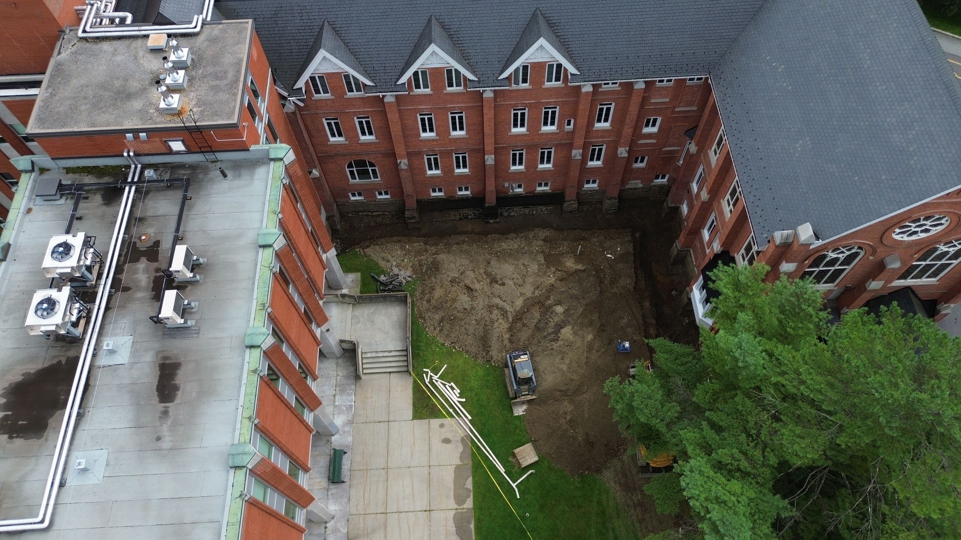 Vue aérienne d'un chantier de construction entre des bâtiments en briques rouges, un sol en terre et des arbres verts.