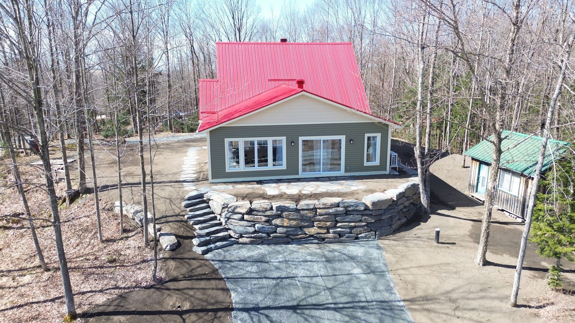 Cabane avec toit rouge et revêtement gris, entourée d'arbres, mur de soutènement en pierre et allée en gravier.