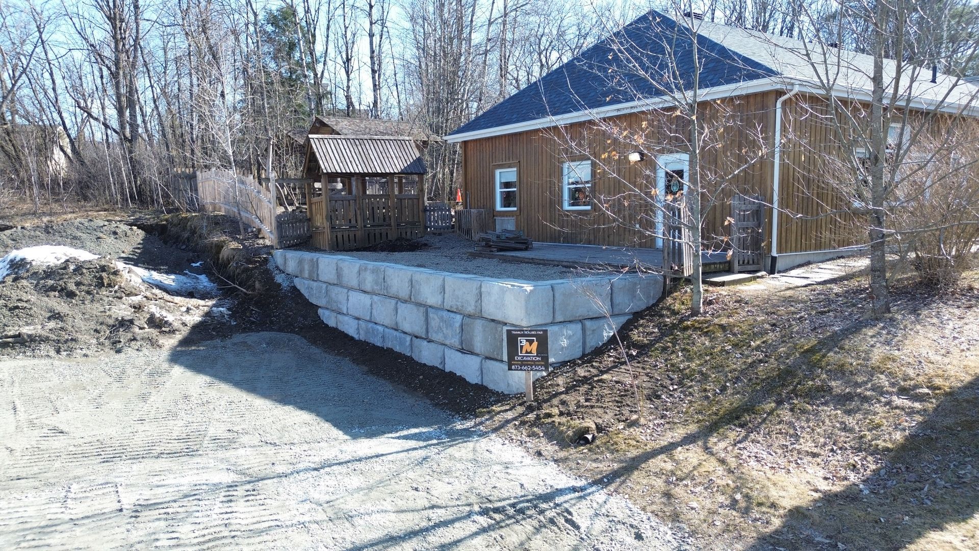 Une maison avec un mur de soutènement en blocs gris, construite sur une colline. Neige présente, journée ensoleillée.