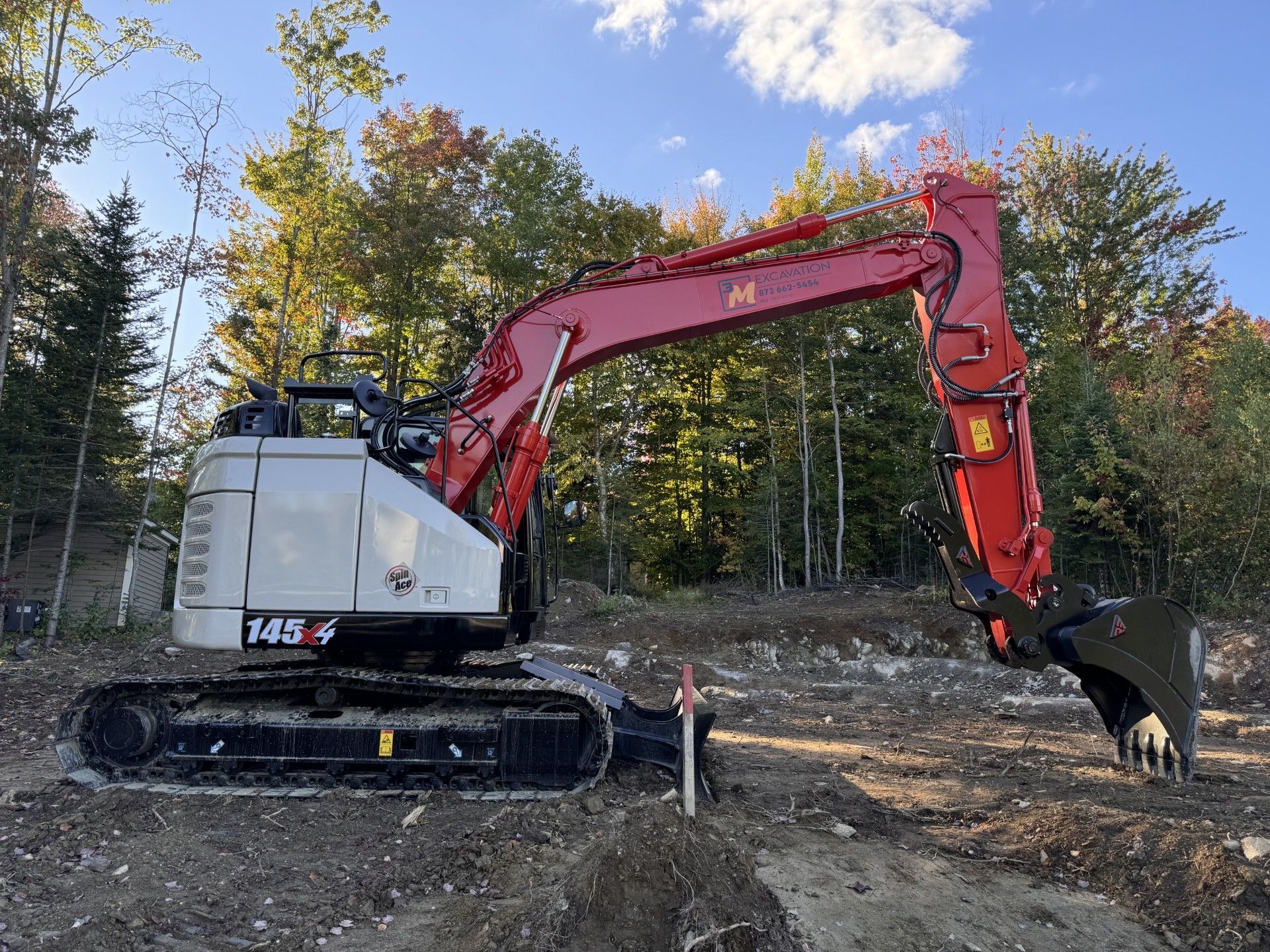 Excavatrice rouge et blanche sur un chantier, bras tendu, avec des arbres en arrière-plan sous un ciel bleu.