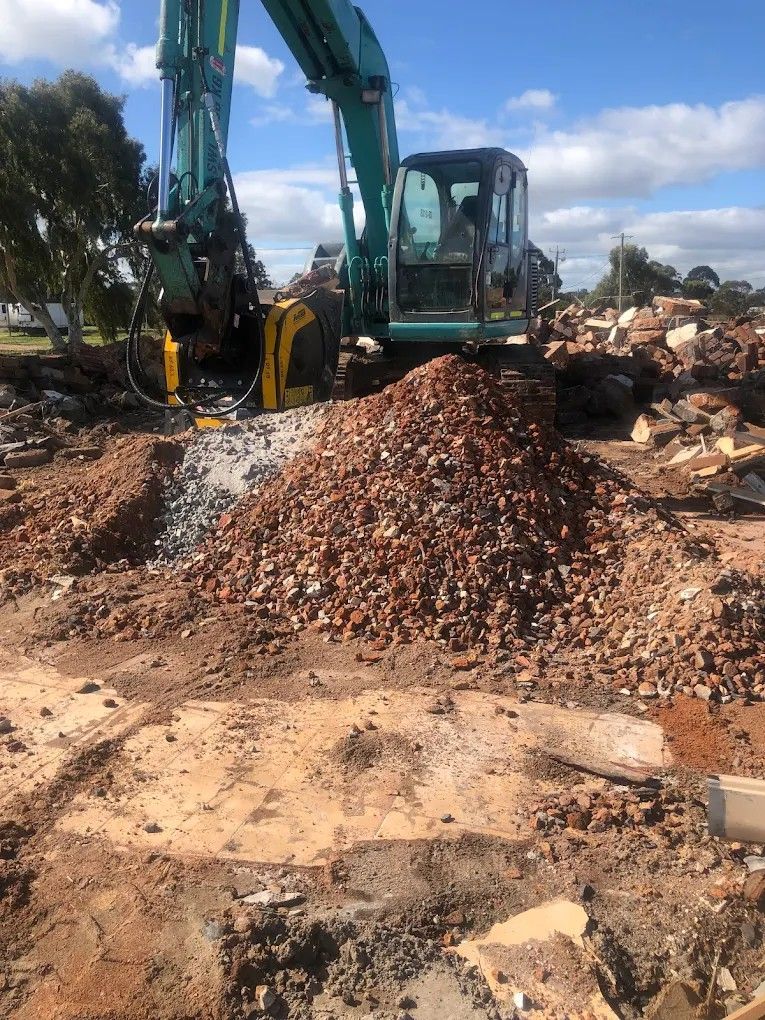 Yellow excavator demolishing a brick structure, with rubble piled in the foreground under a blue sky