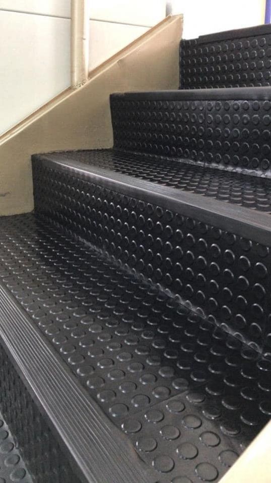 Escaleras recubiertas de caucho negro con un patrón redondeado, que ascienden junto a una pared blanca.