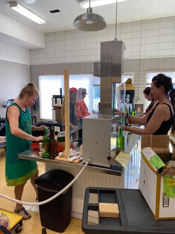 People bottling beverages in a brightly lit kitchen. One woman in green, two in black, and others in the background.