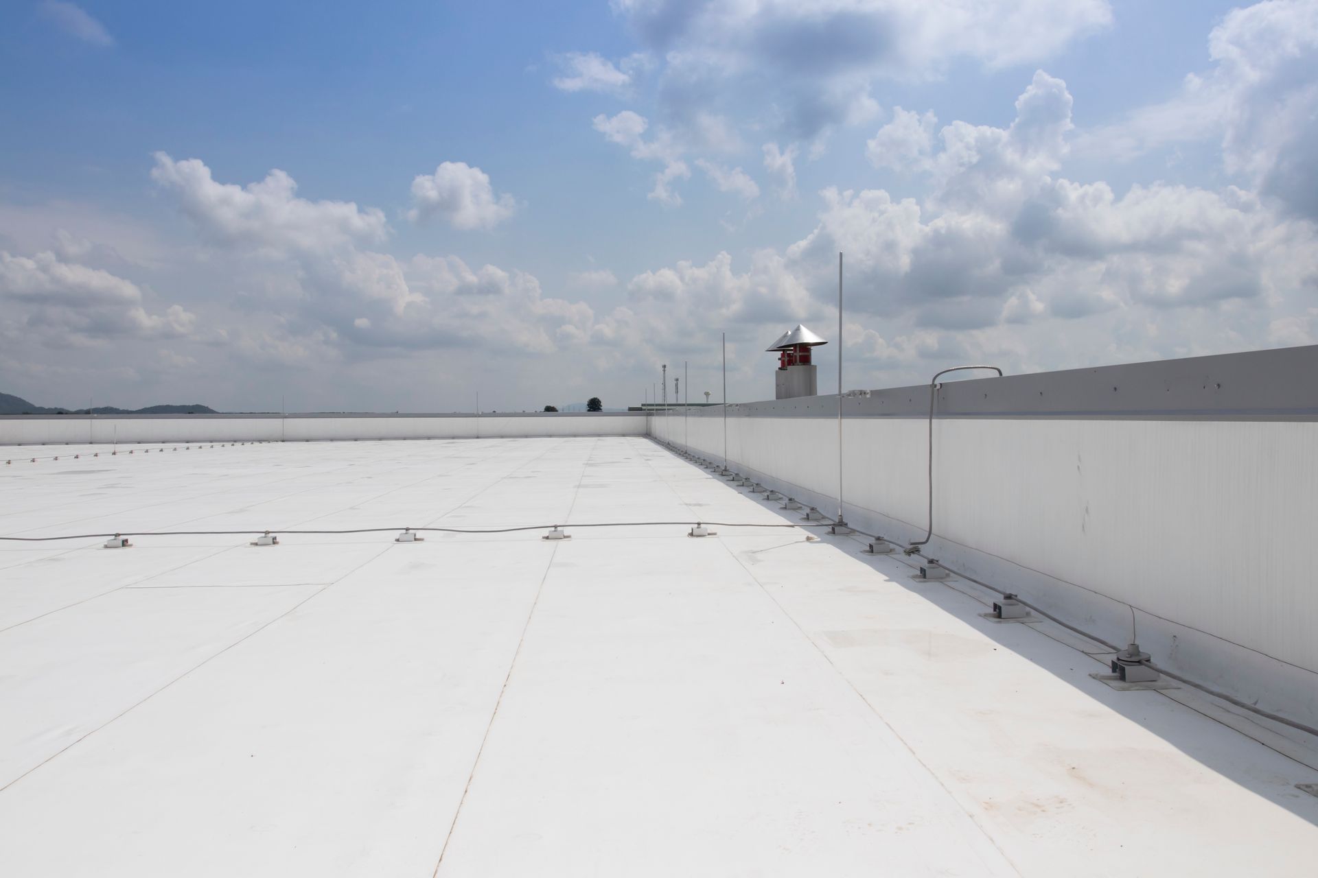 A flat white rooftop under a cloudy blue sky, featuring a parapet wall with an installed lightning protection system.