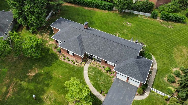 An aerial view of a brick house with a dark gray shingled roof, a paved driveway, and a spacious green lawn.