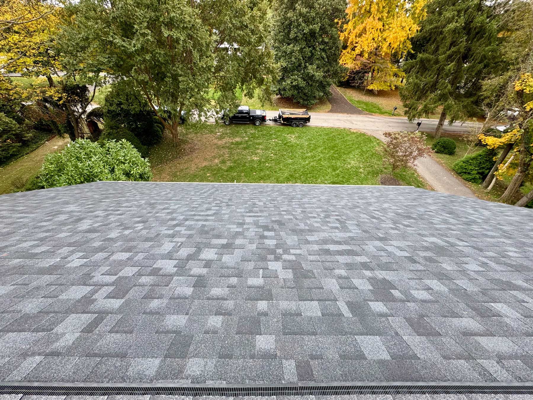 A view from a roof looking down at a grassy yard, driveway with two parked vehicles, and surrounding autumn trees.