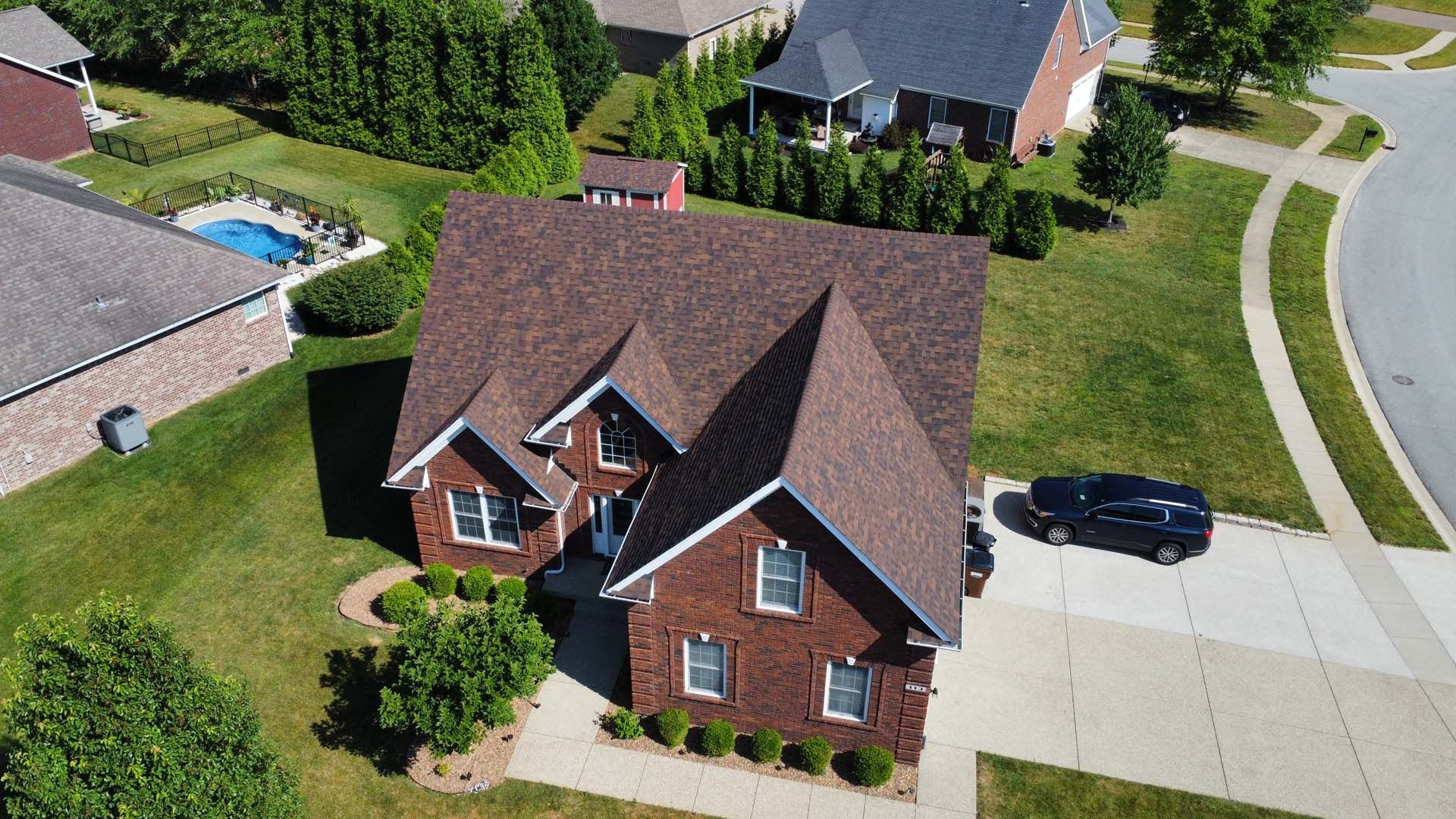 An aerial view of a brick house with a brown shingled roof, a driveway with a dark car, and surrounding green lawns.
