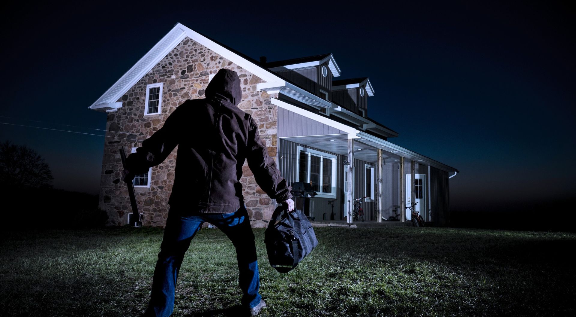 Person in hoodie exiting a house at night, carrying a bag, and holding a weapon.