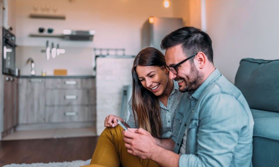 Couple smiling, looking at wi-fi security and home security, sitting on floor in modern house.