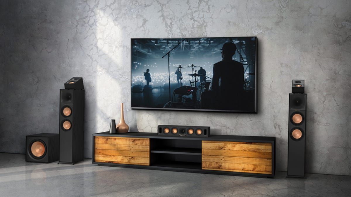 A home entertainment setup with TV, soundbar, speakers, and subwoofer against a textured gray wall.