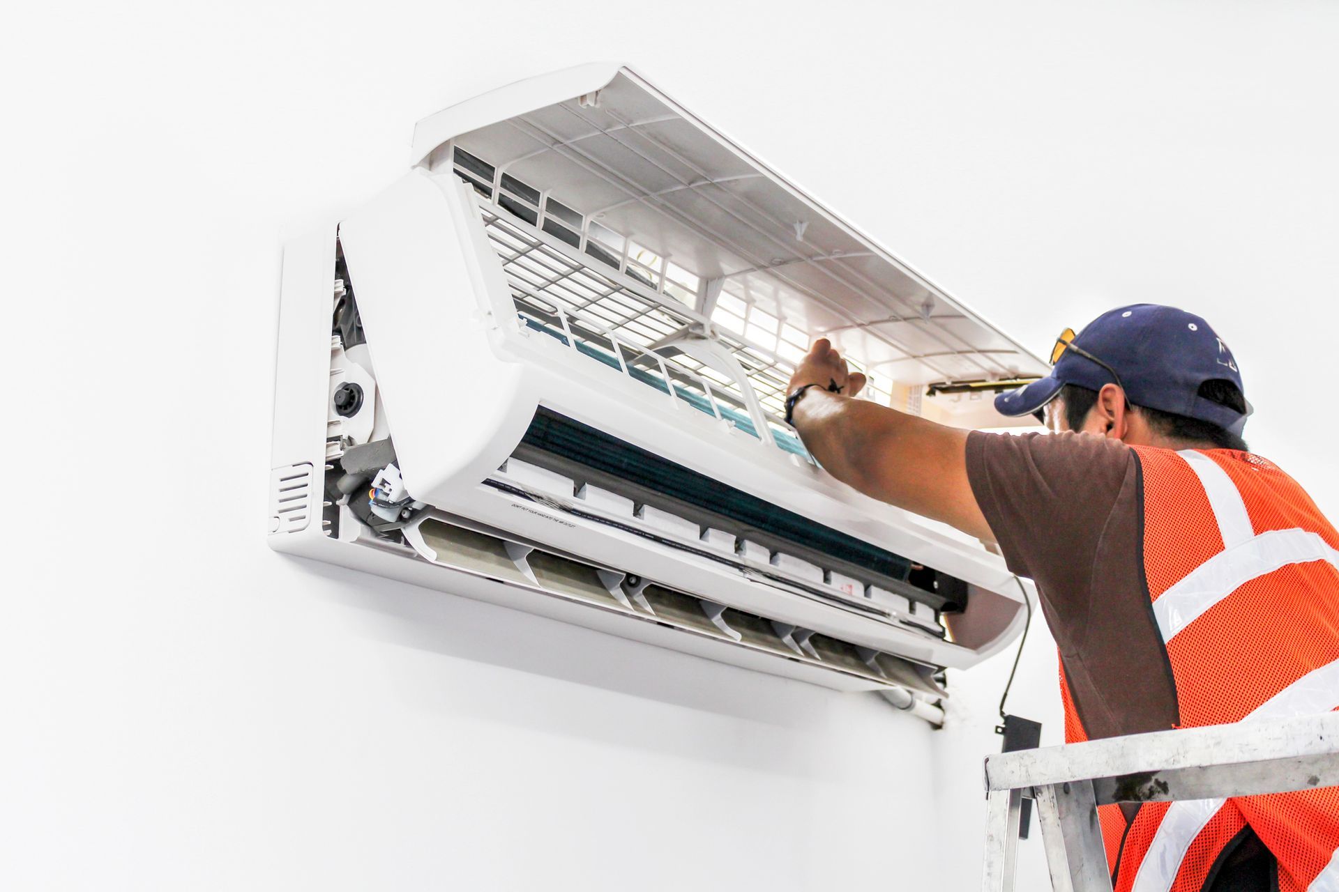 A technician in a high-visibility vest stands on a ladder to repair a wall-mounted air conditioner.