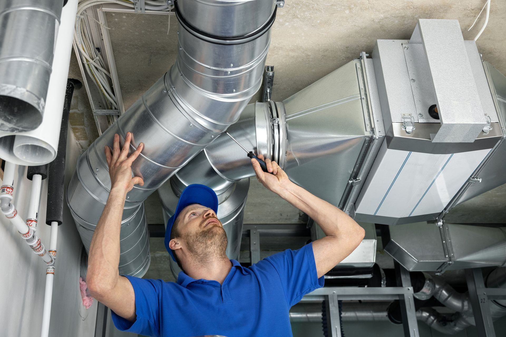 Technician in blue shirt inspecting silver HVAC ductwork overhead in an industrial space