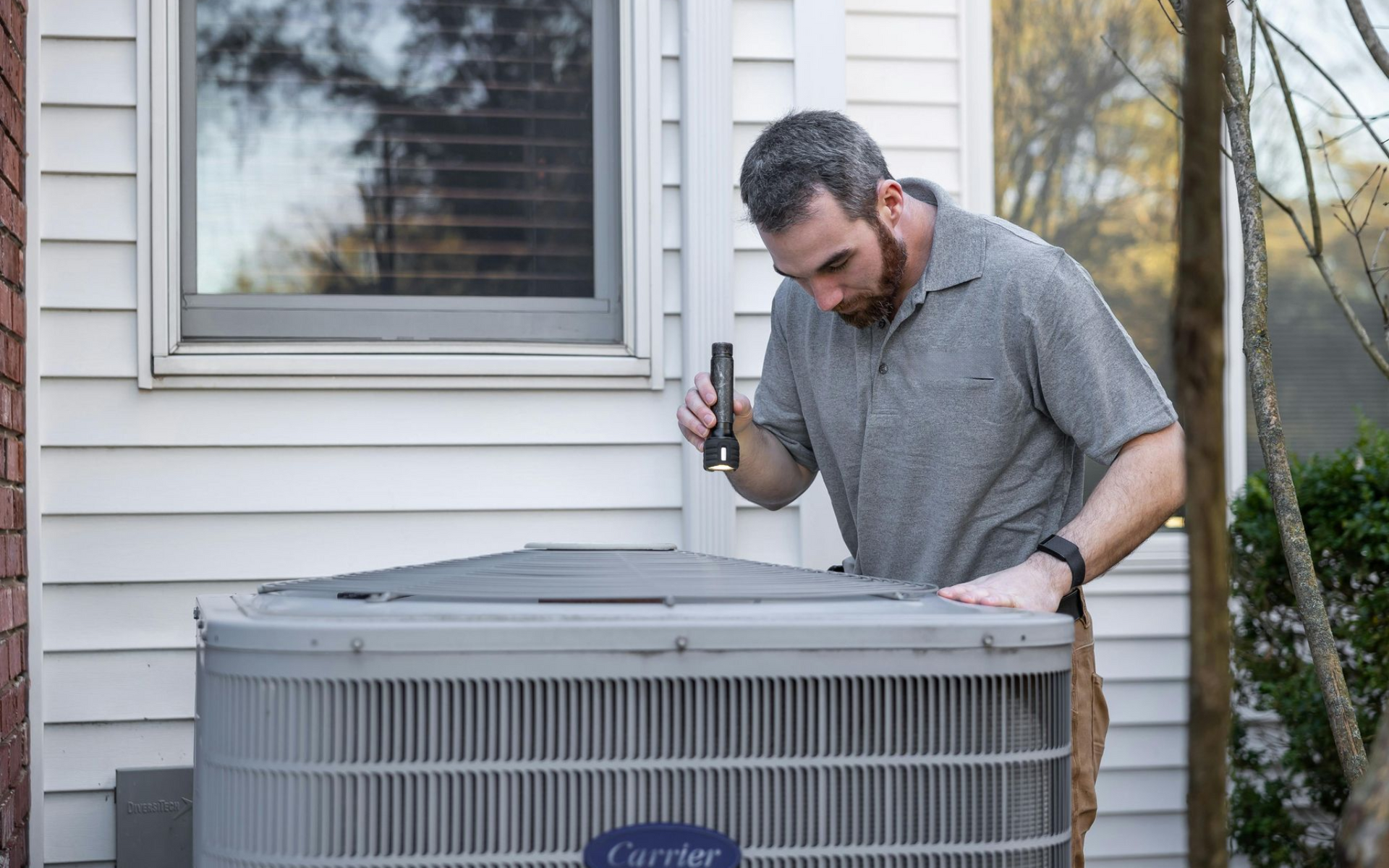 Man inspecting an air conditioning unit with a flashlight outside a house.