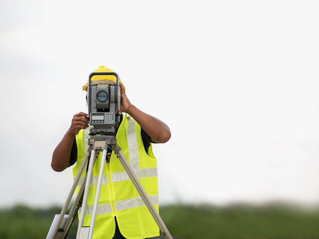 A man in a yellow vest is using a theodolite on a tripod.