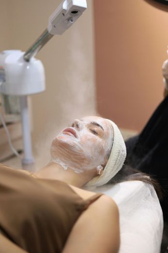 Woman receiving facial steam treatment at a spa, with a mask on her face and head band.