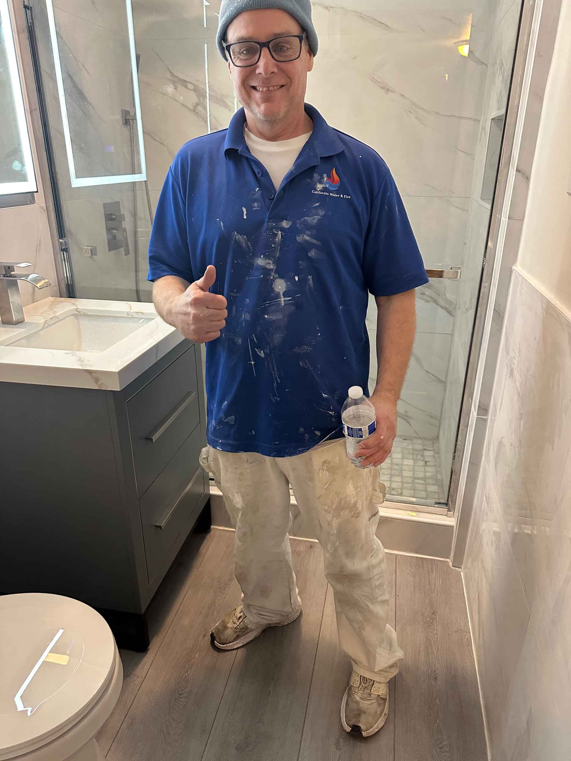 A man in a blue shirt and white pants is standing in a bathroom giving a thumbs up.