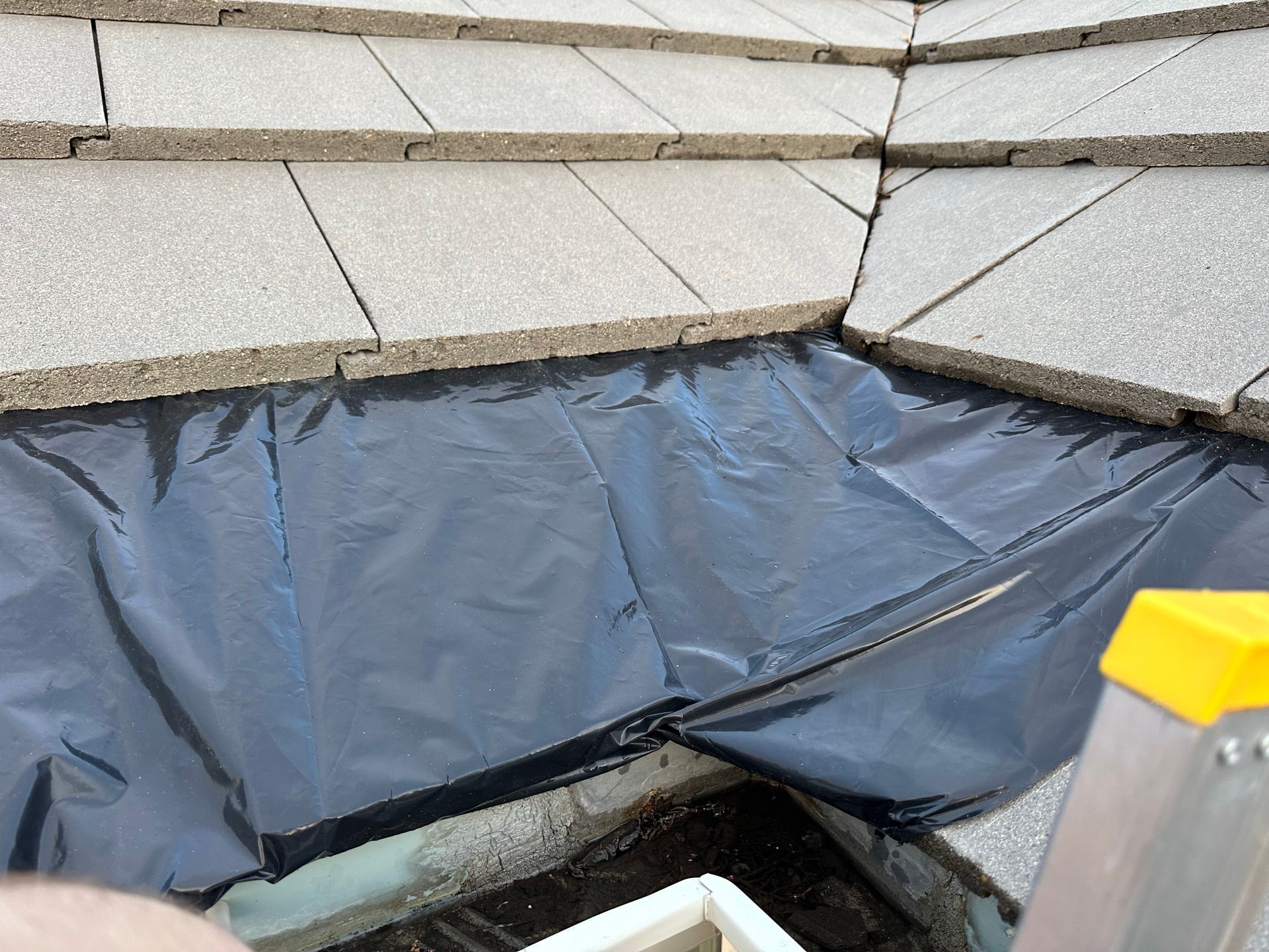 A person is standing on a ladder next to a piece of plastic on a roof.