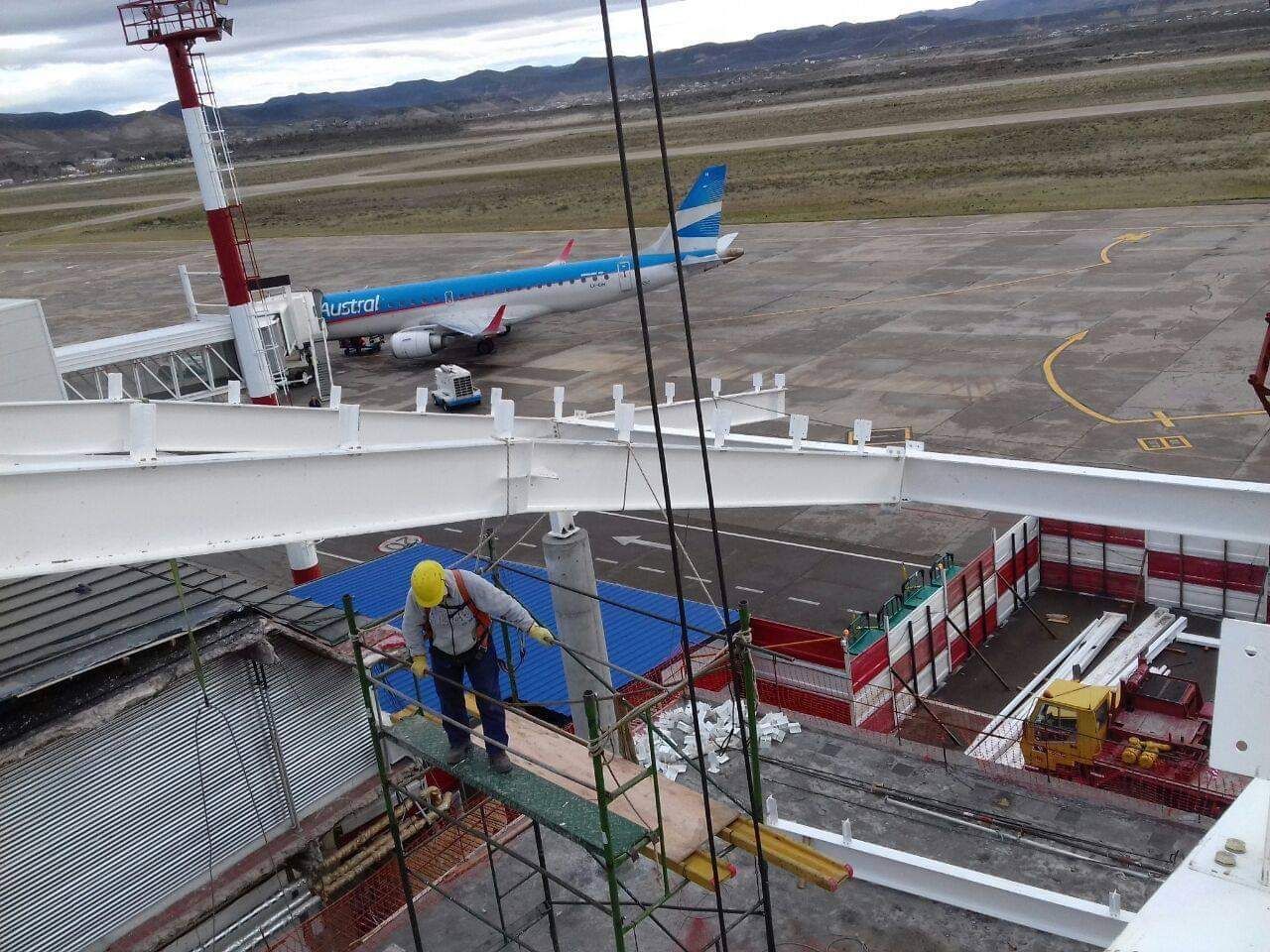 Un hombre está de pie sobre un andamio en un aeropuerto con un avión al fondo.