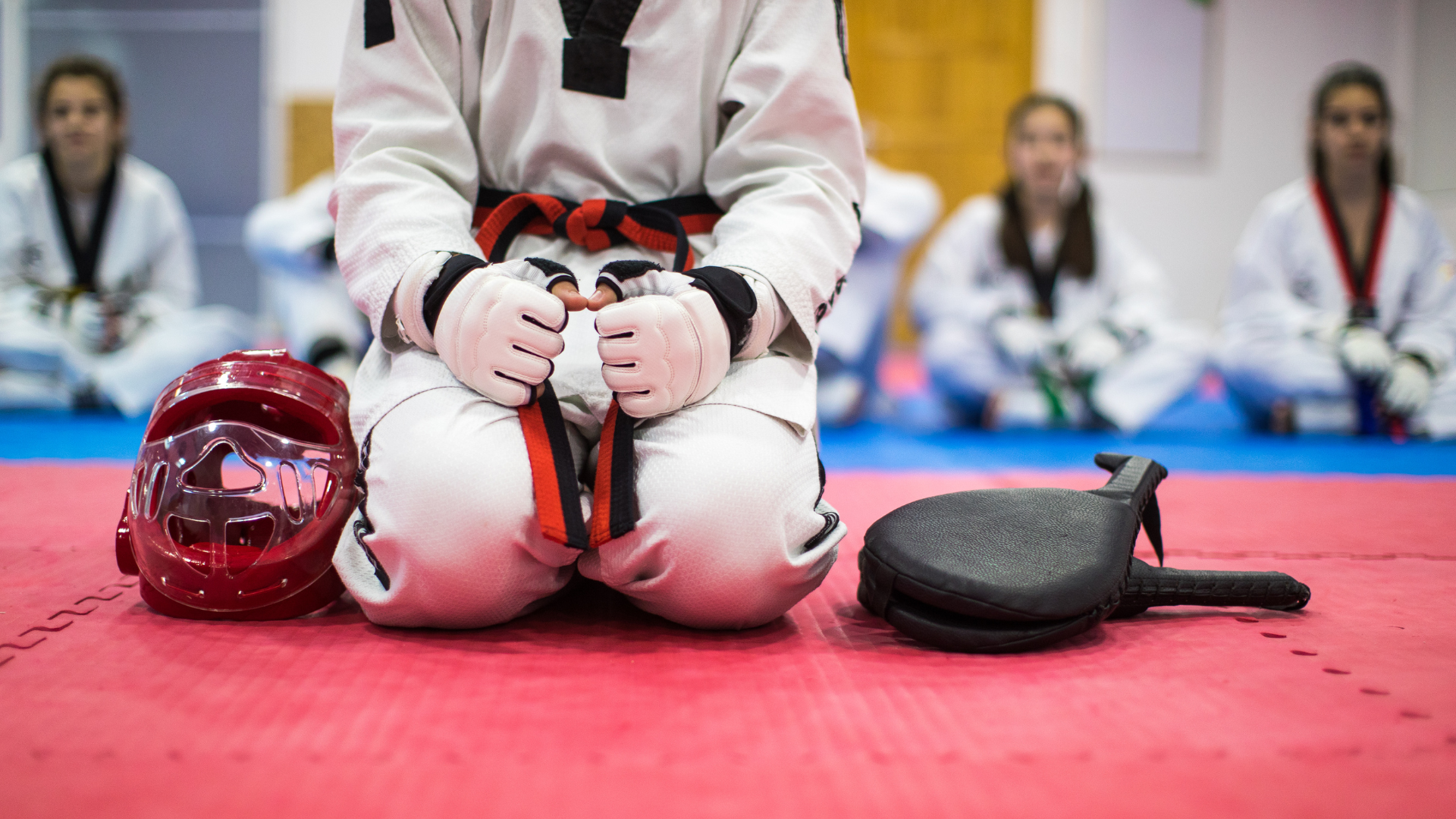 A person in a taekwondo uniform is sitting on the floor next to a helmet and a paddle.