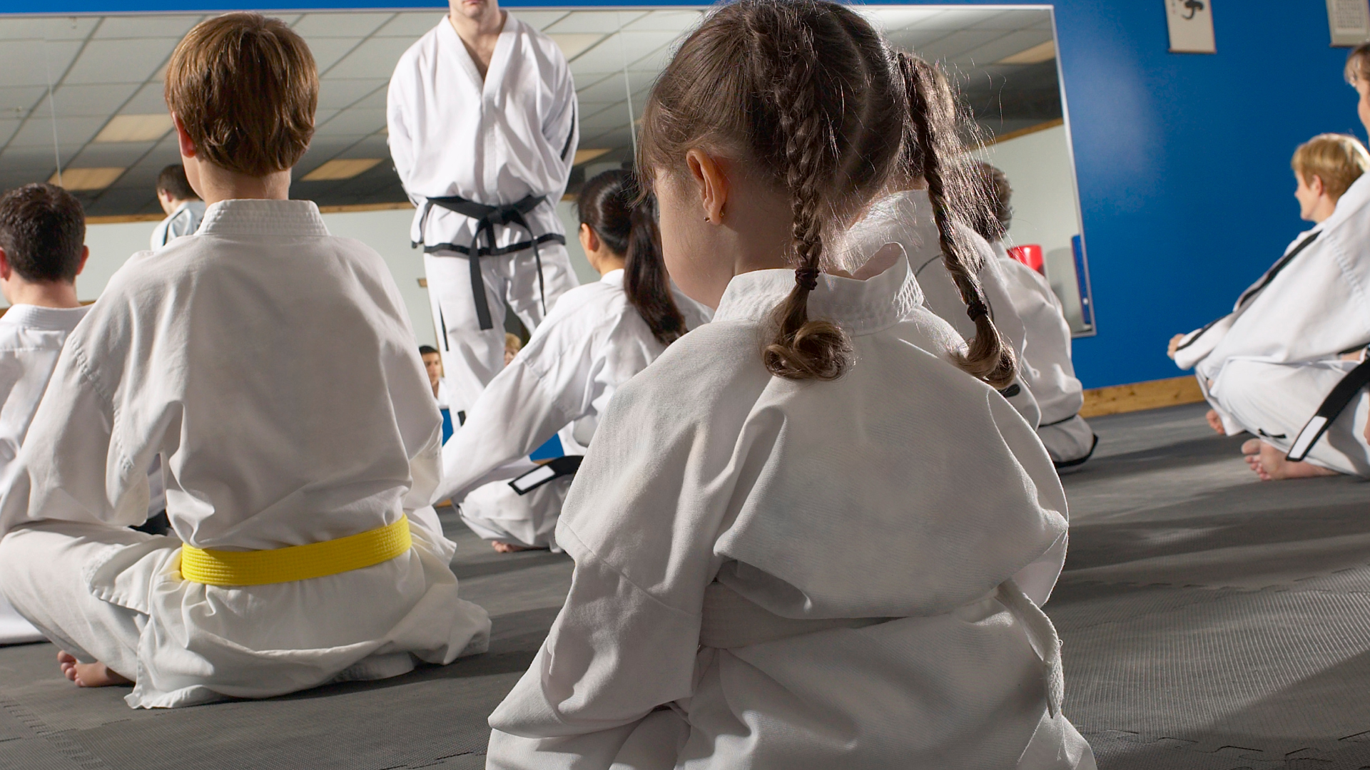 A group of children in karate uniforms are sitting on the floor in front of a mirror.