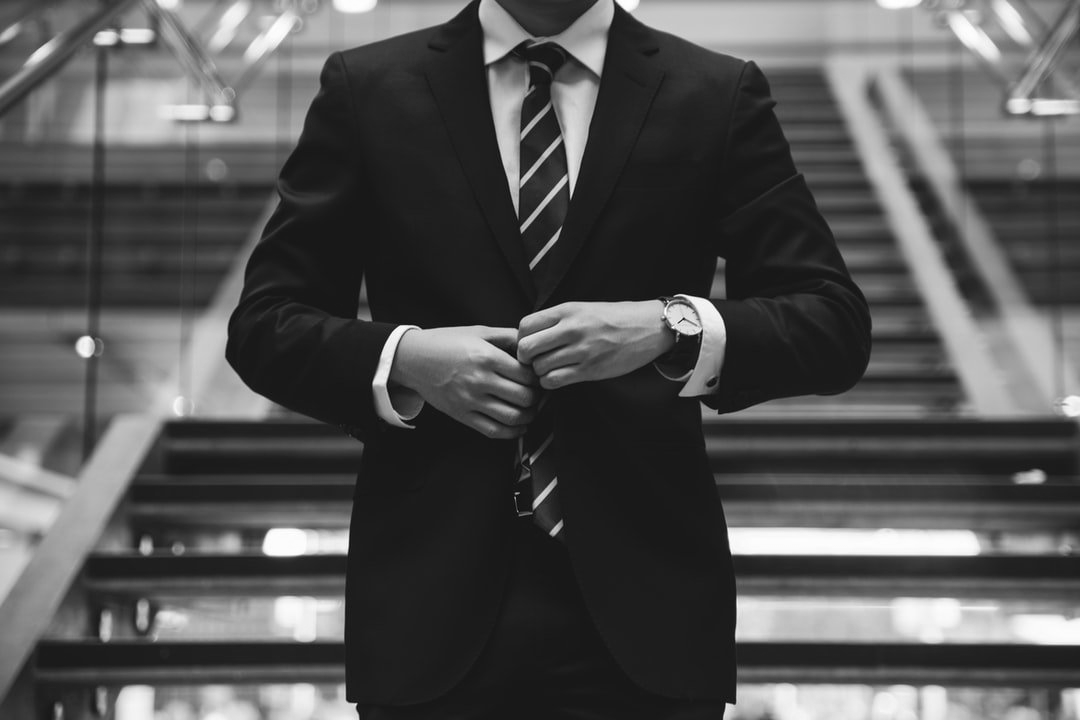Man in a suit buttoning his jacket, standing on stairs with a glass railing.