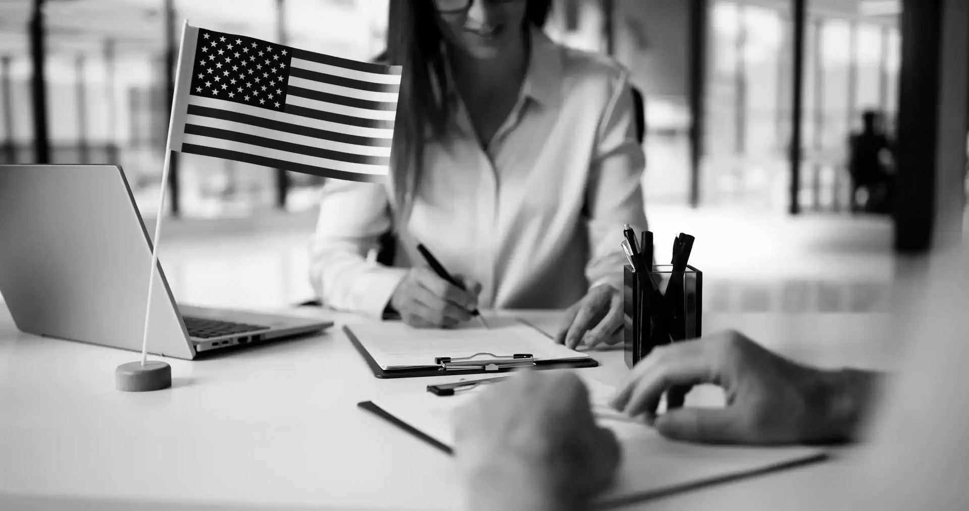 Woman writing at desk with small US flag and laptop, possibly in an office.