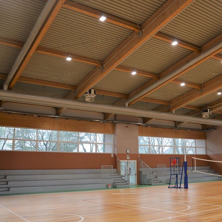 Gymzaalinterieur met houten vloeren, tribunes en volleybalnet, onder een hoog plafond voorzien van metalen geperforeerde luchtverdeelkanalen
