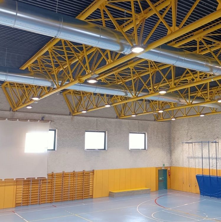 Het interieur van een sportschool met een geel spantenplafond, metalen geperforeerde luchtkanalen en kleine ramen.