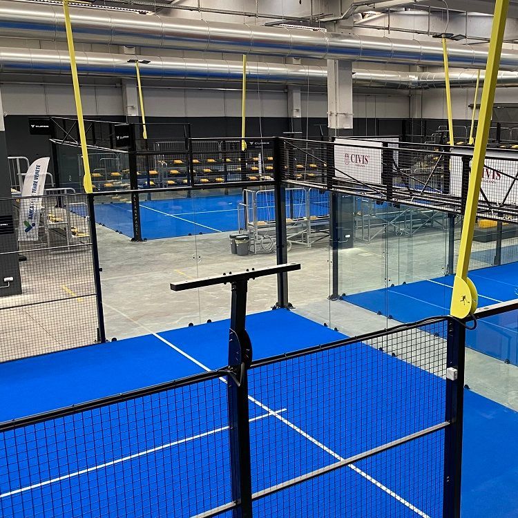 Overdekte padeltennisbanen met blauwe ondergrond en netten, van bovenaf gezien met metalen luchtverdeelkanalen