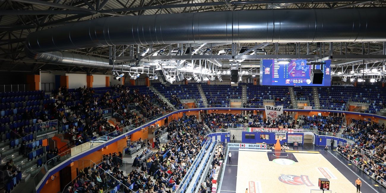 Basketbalarena met volle tribunes. Spelers op het veld; scorebord zichtbaar. Blauwe stoelen met oranje accenten. met micro geperforeerde metalen luchtverdeelkanalen