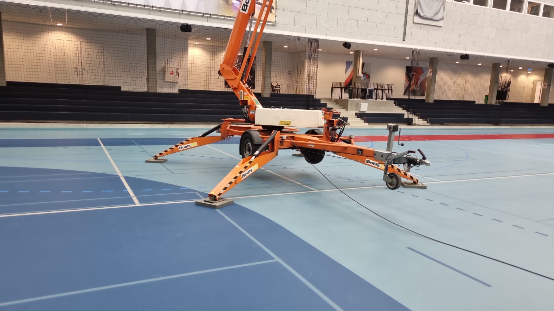 Een oranje hoogwerker voor werk aan de airsocks/luchtverdeelslangen in een sporthal.