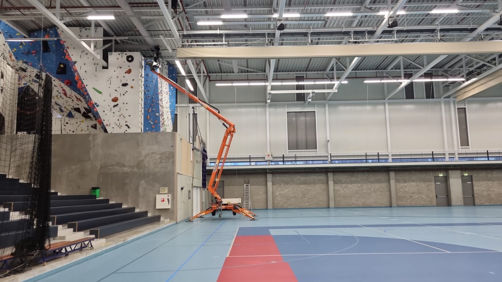 Een hoogwerker is aan het werk aan airsocks/luchtverdeelslangen hangende aan het plafond van een grote sporthalruimte.