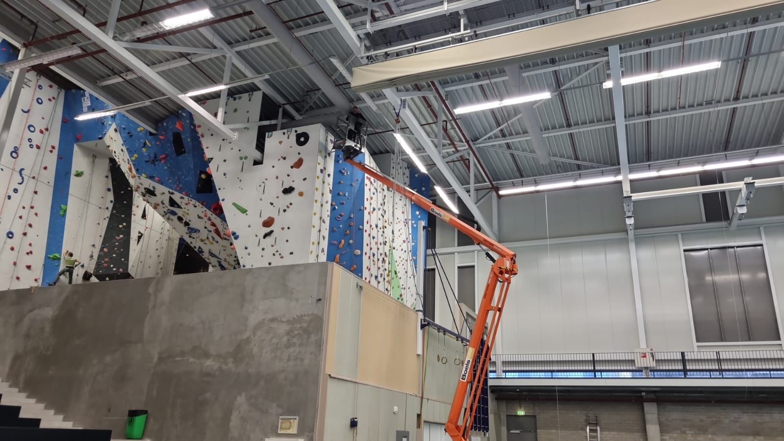 Een hoogwerker voor het onderhoud van een airsock/luchtverdeelslang boven een klimwand in een sporthal.