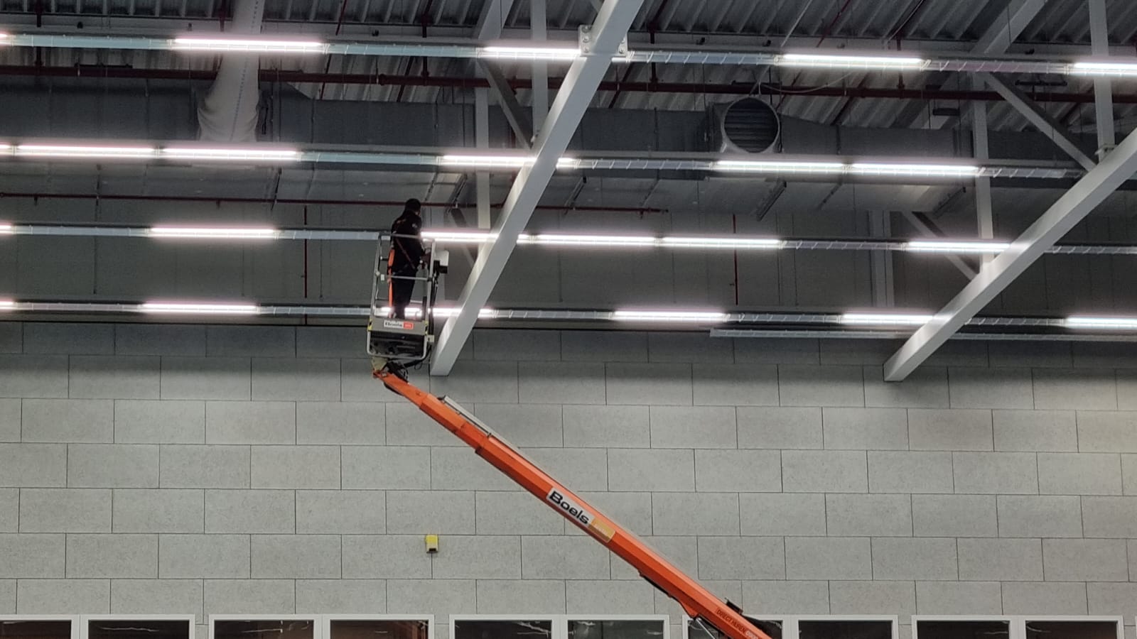 Een man staat op een hoogwerker en werkt aan airsock/luchtverdeelslang aan het plafond van een gebouw.
