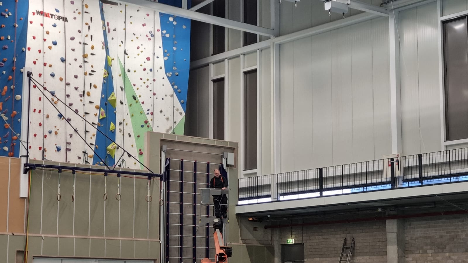 Een man in een hoogwerker voor het onderhouden van airsocks/luchtverdeelslangen boven een klimwand in een sportschool.