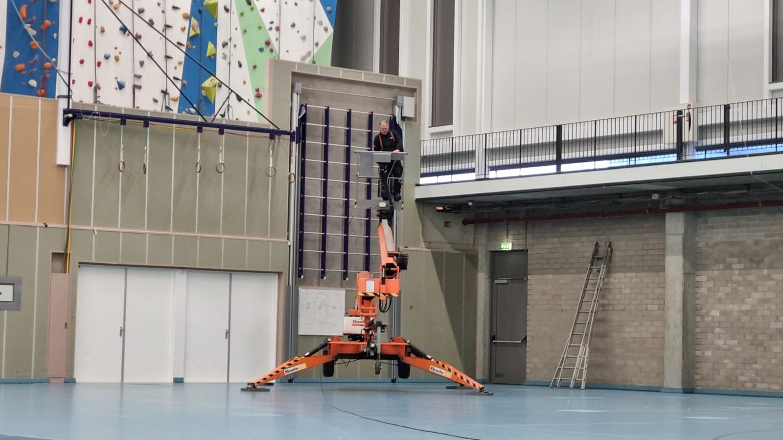 Een man staat op een hoogwerker in een sporthal.