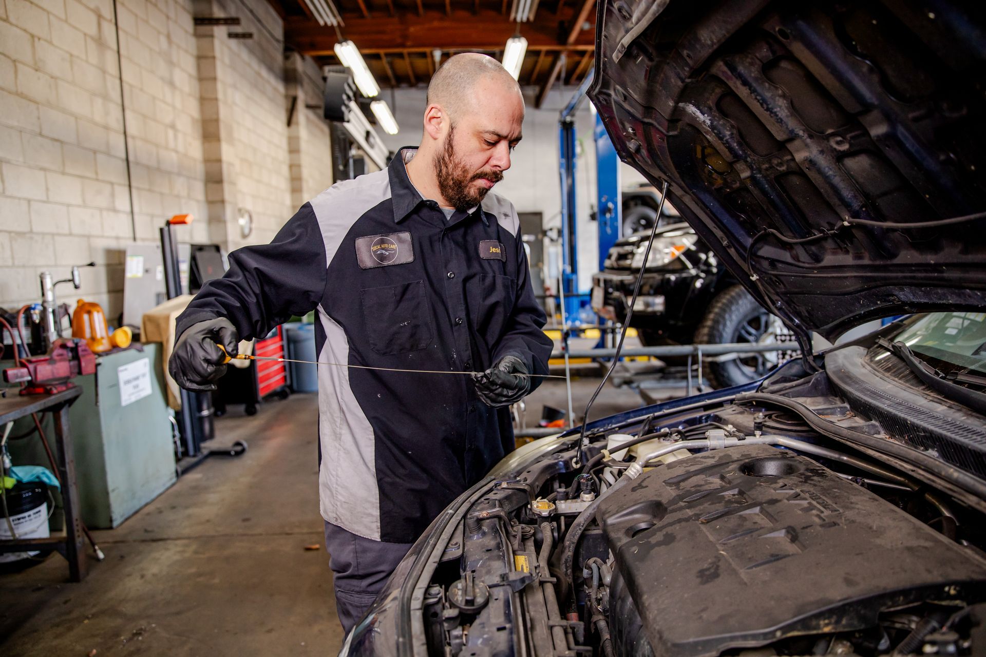 A mechanic in a black and grey uniform checks the oil level using a dipstick in a car with its hood open at a workshop.