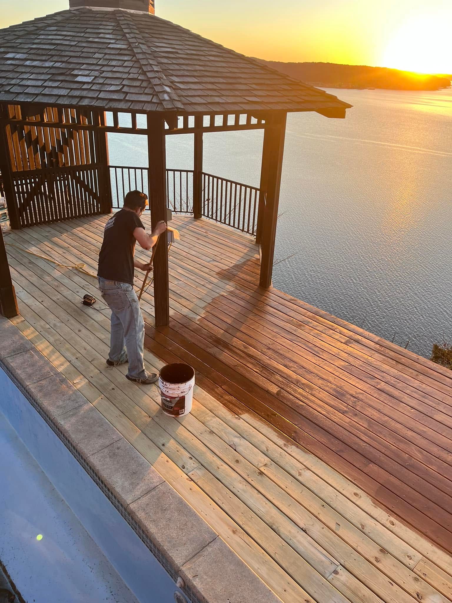 Man staining wooden deck of a gazebo overlooking water at sunset.