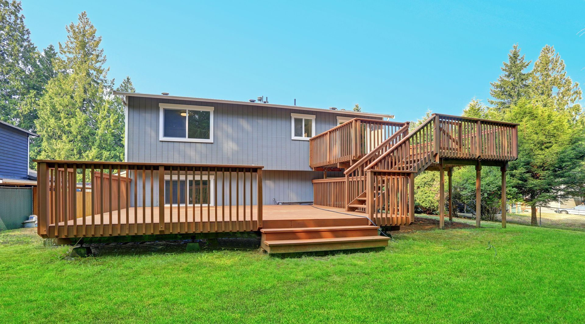A wooden deck extends from a two-story gray house, with stairs, in a grassy backyard.