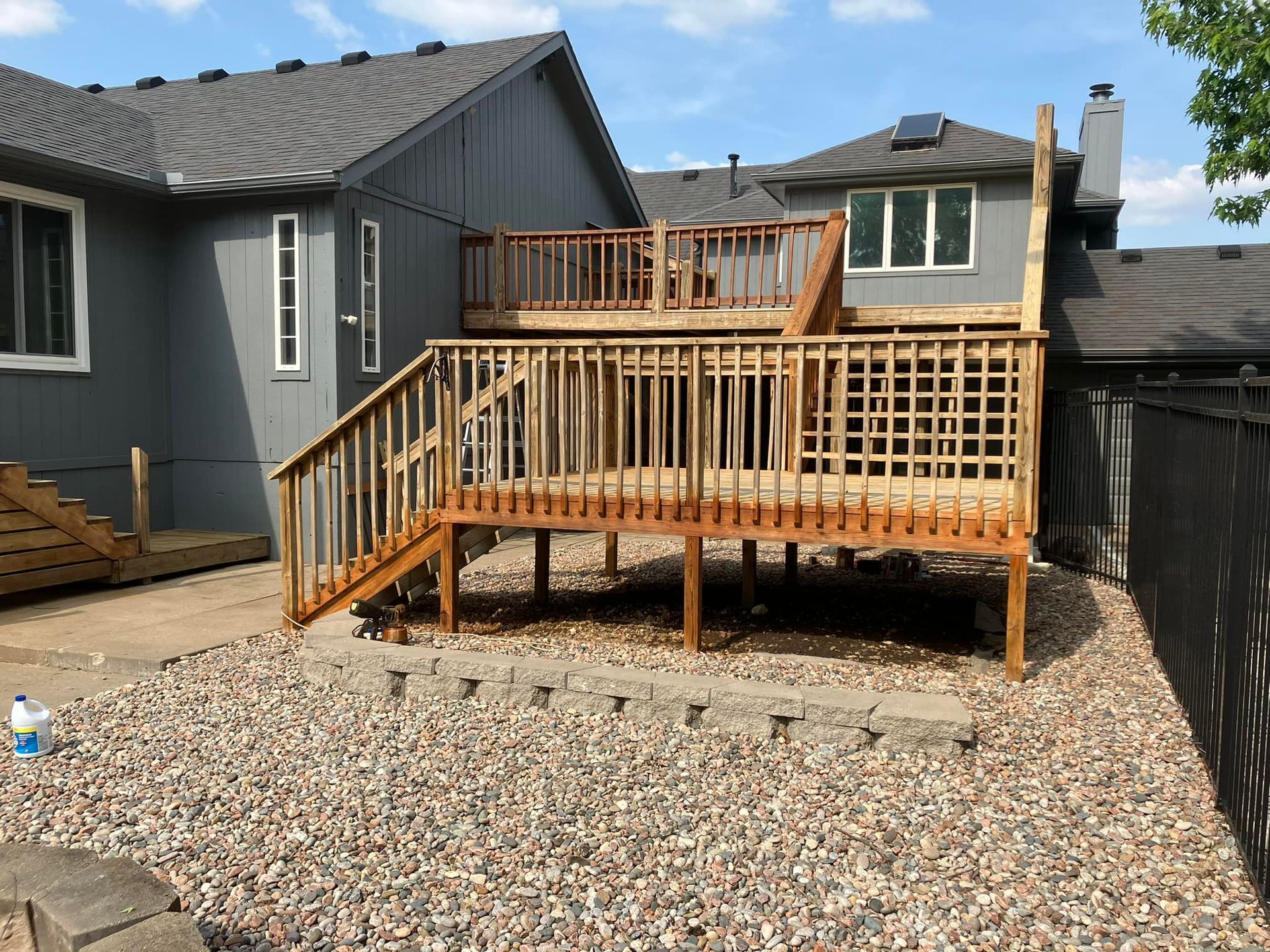 Two-level wooden deck attached to a grey house, over gravel and a low retaining wall, under a blue sky.