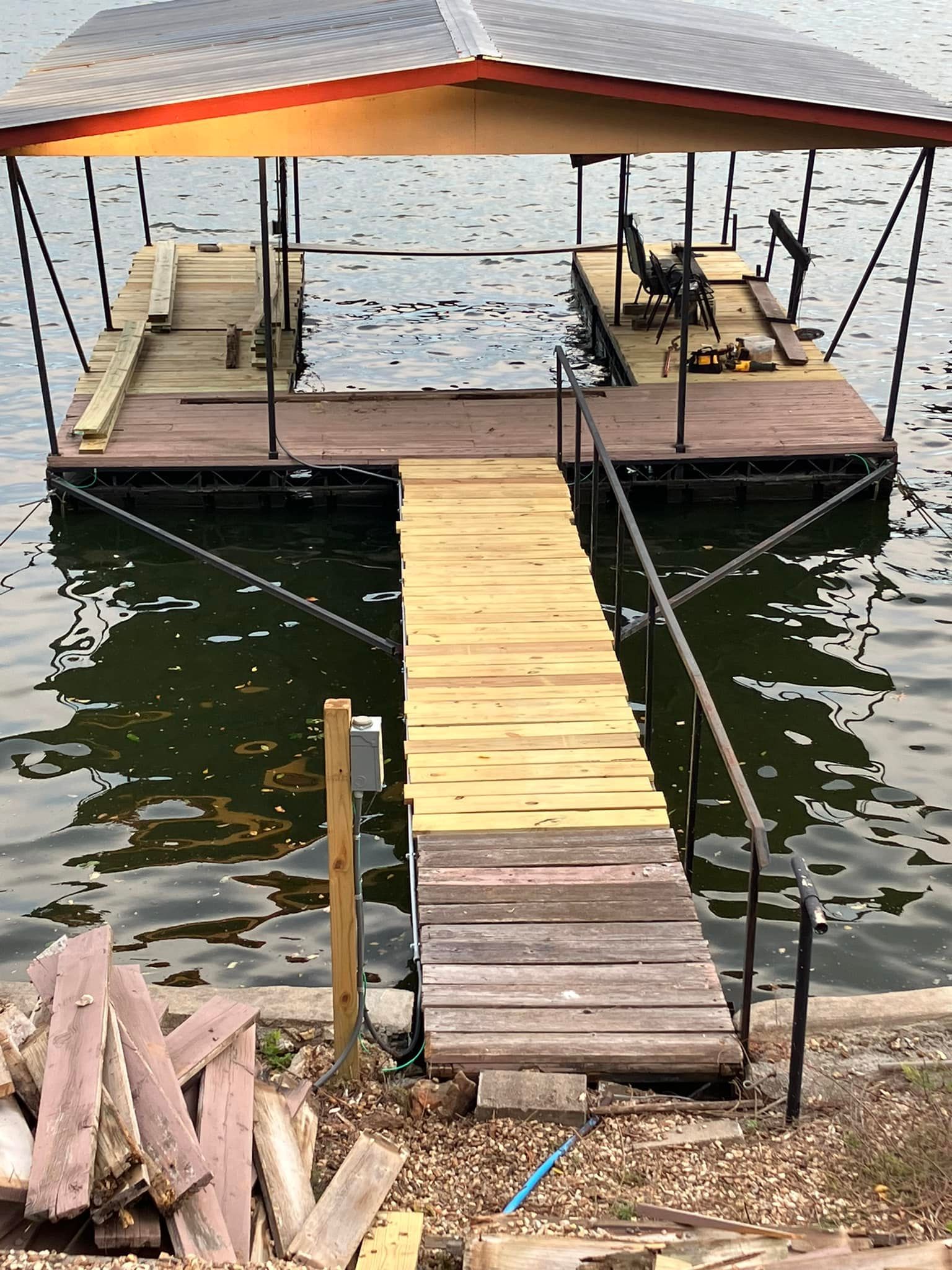 Dock extending into water with a covered boat slip, wooden planks, black railing, and scrap wood on shore.