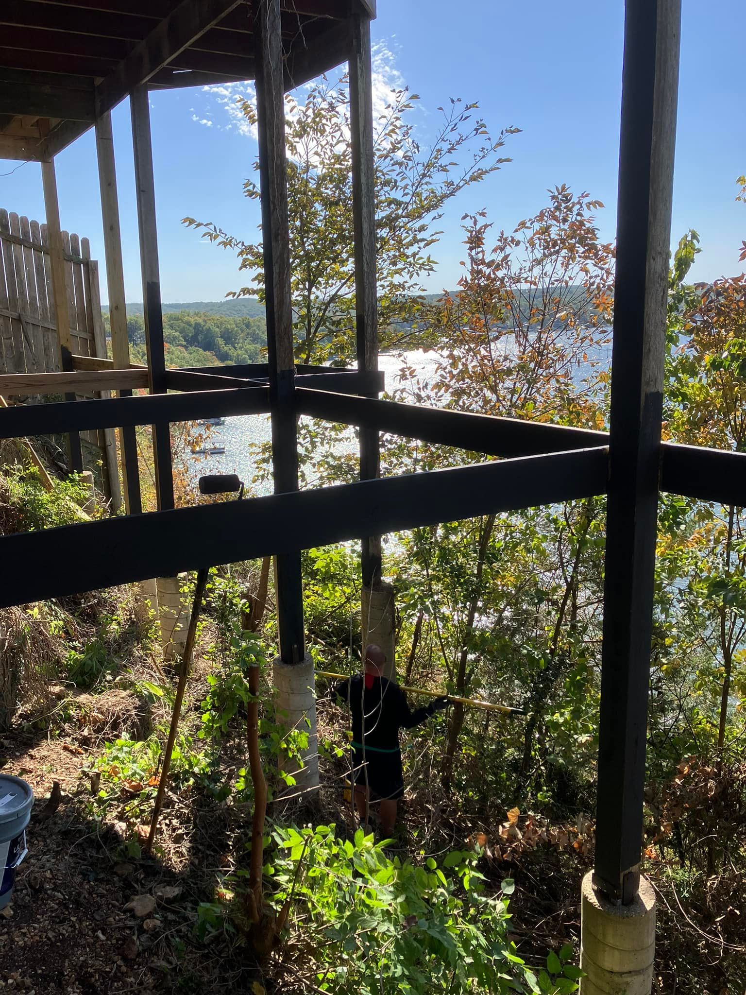 Person standing under a deck overlooking water, surrounded by trees. Sunny day.