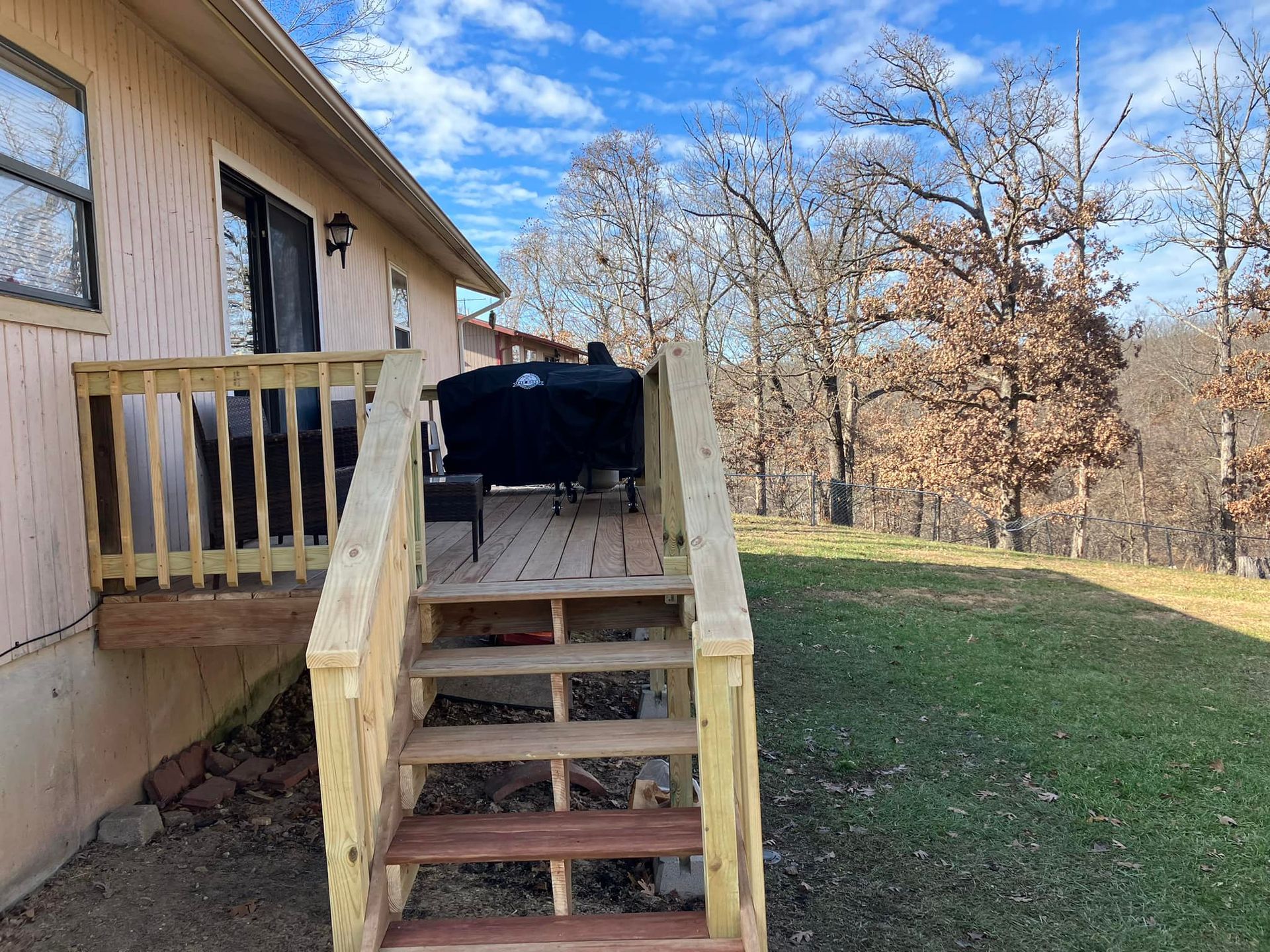 Wooden deck with stairs leading to a house with a grassy yard and trees under a blue sky.