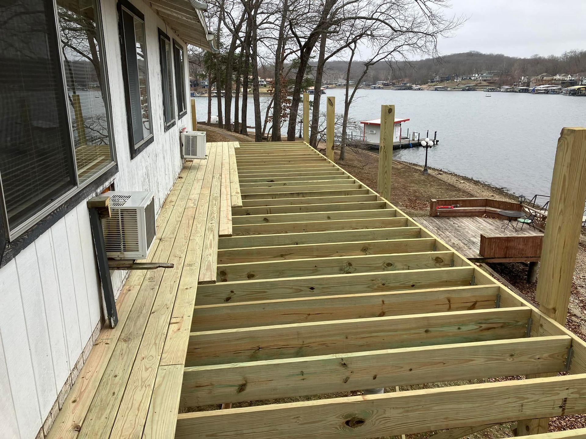 Deck construction: Wooden beams and partially laid planks next to a white house, with a lake in the background.