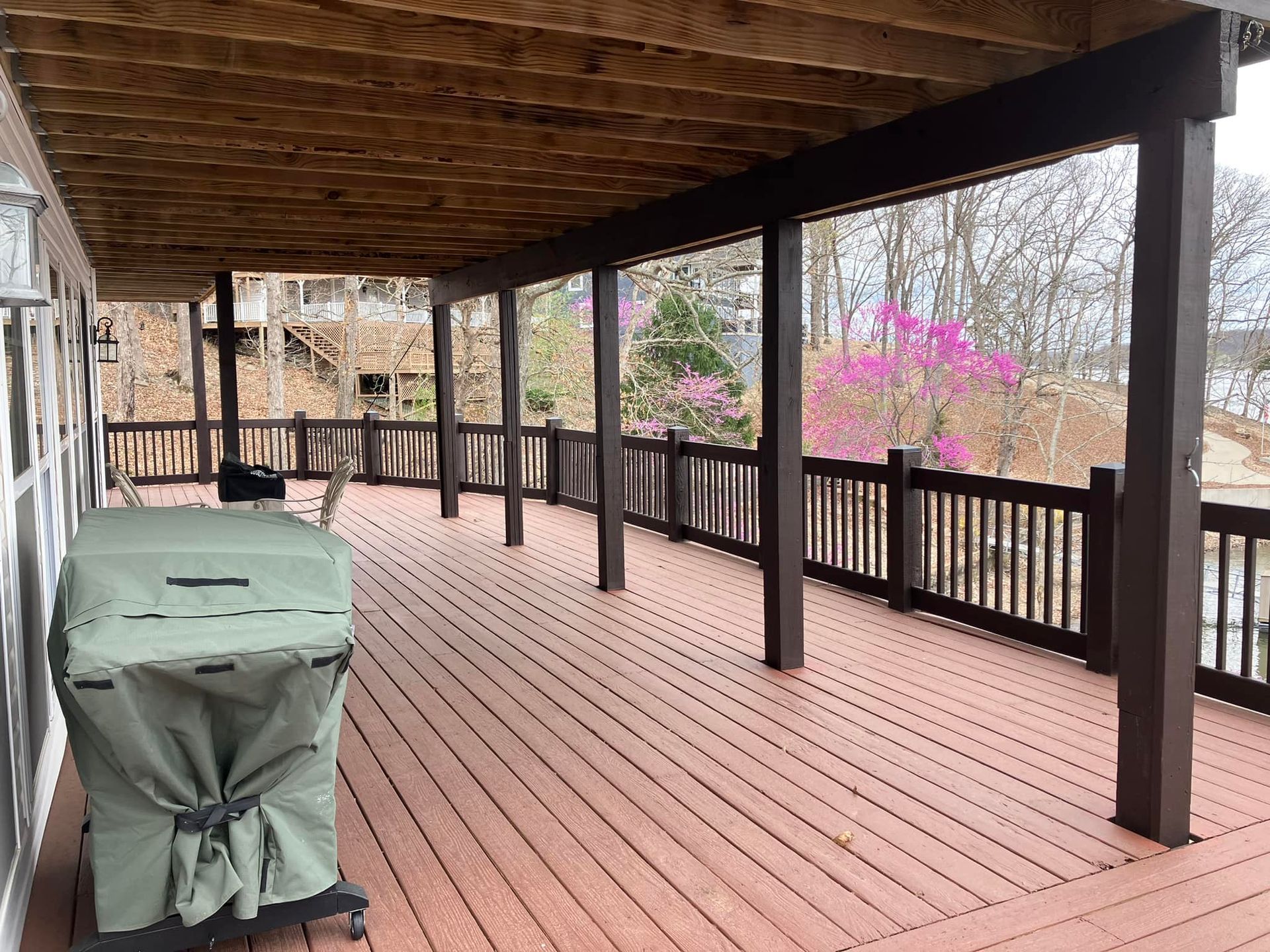 Covered wooden deck with grill; dark posts, railings; trees and pink blooms in the background.