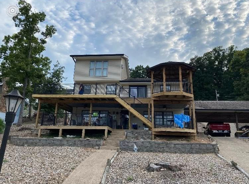 Two-story house on rocky shoreline with decks, gazebo, car port, and blue sky.
