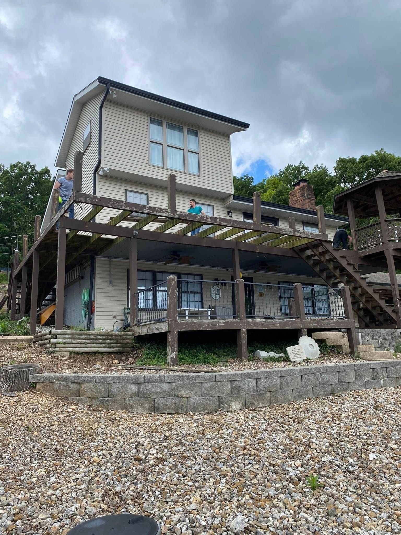 Two-story lakeside house with brown pergola, deck, and rocky shore under overcast sky.