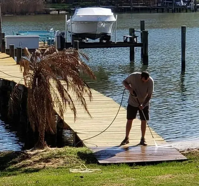 Man pressure washing a wooden dock next to a body of water. Boat and dock posts in the background.