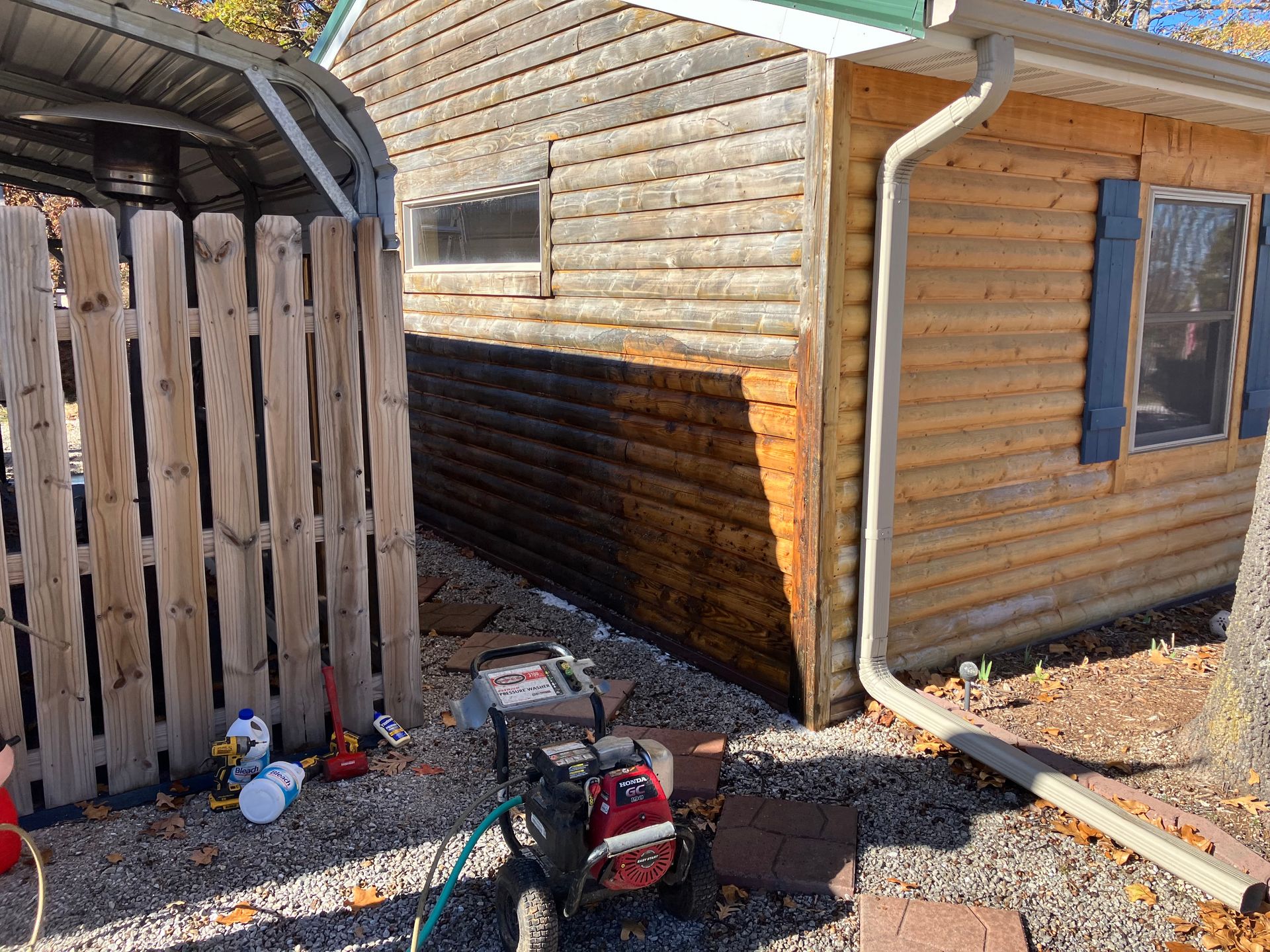 A power washer cleans a section of a log cabin. Brown wood contrasted by a darker, cleaned section.